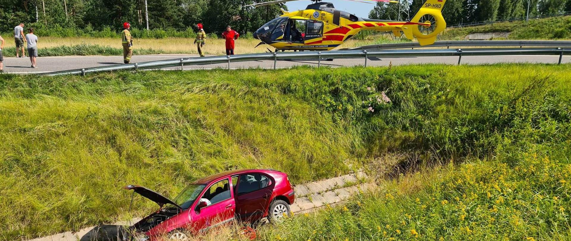 Zdjęcie obrazuje samochód osobowy, który wpadł do głębokiego rowu. Na drodze nieopodal pojazdu stoi helikopter LPR, ratownik medyczny oraz dwóch strażaków. W tle drzewa i słup energetyczny.