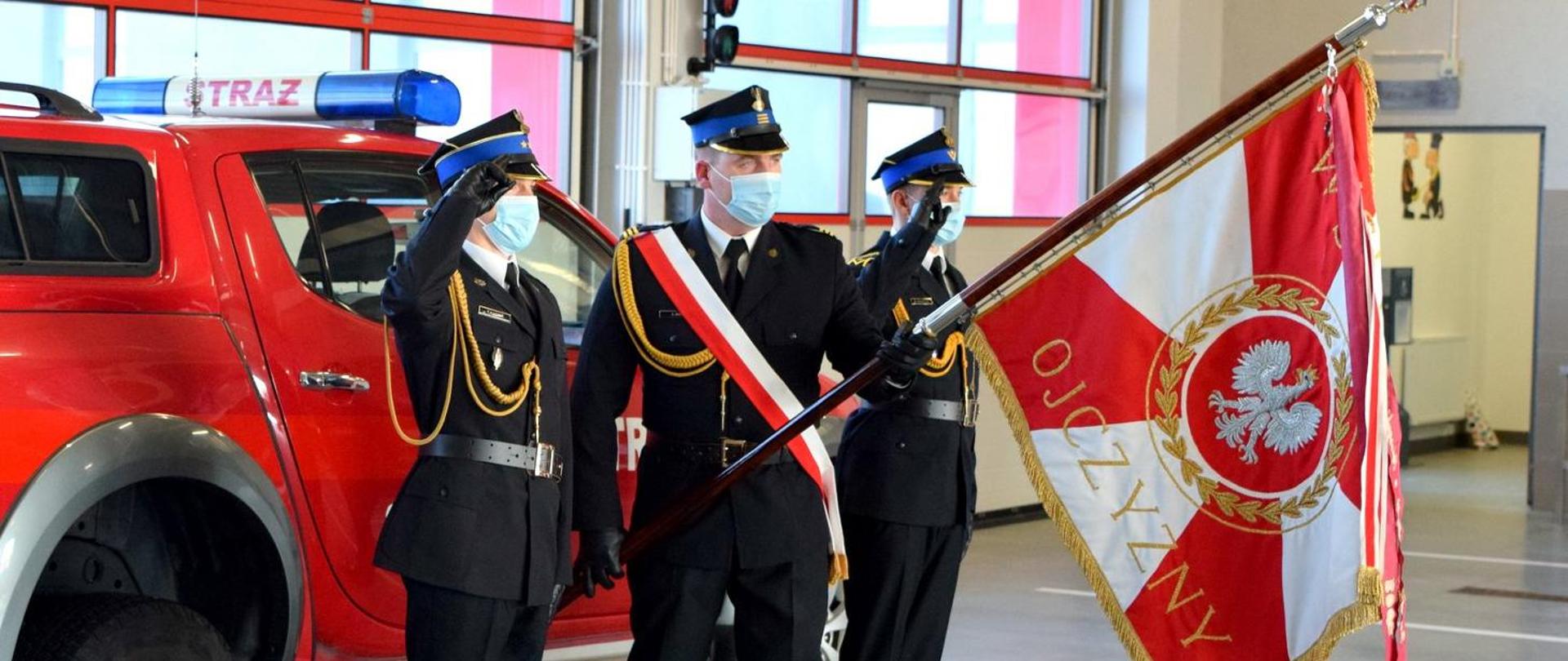 Zdjęcie zrobione w pomieszczeniu. Na zdjęciu widać poczet sztandarowy składający się z trzech strażaków, ubranych w mundury wyjściowe. Strażak ustawiony w środku przepasany jest biało-czerwona szarfą i trzyma pochylony sztandar. Obok niego funkcjonariusze salutują. Za strażakami widać samochód pożarniczy.