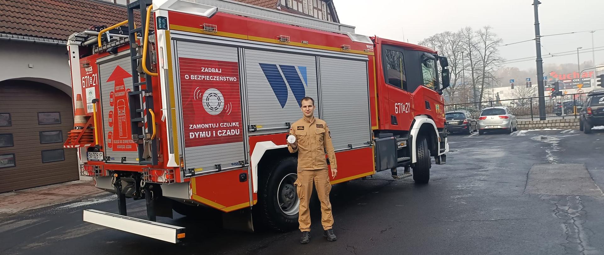 Na zdjęciu widoczny jest strażak trzymający w dłoni i prezentujący przed sobą czujkę dymu. Za nim stoi samochód pożarniczym. W tle budynek komendy. Pogoda pochmurna.