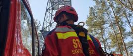 Ratownik w kombinezonie wysokościowym i kasku. Ma na sobie sprzęt wysokościowy. Na drugim planie wieża obserwacyjna Lasów Państwowych. Niebo niebieskie. W tle drzewa iglaste. Po lewej pojazd straży pożarnej w kolorze czerwonym.