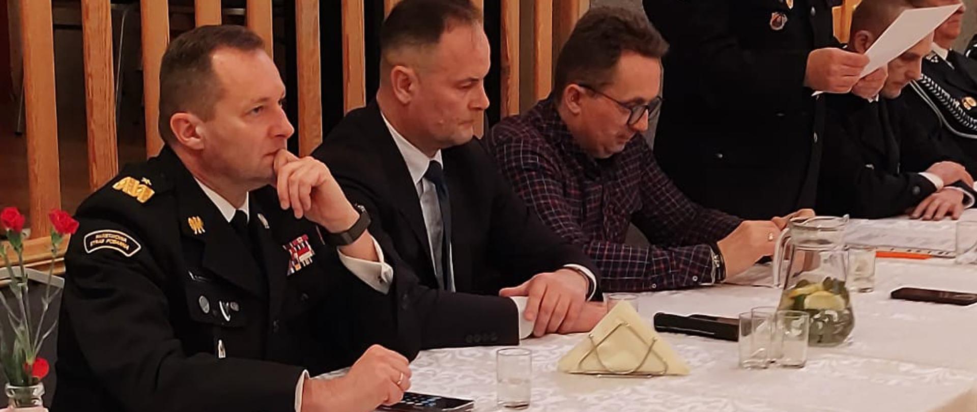 Mężczyźni siedzący przy stole, jeden z nich w mundurze Ochotniczej Straży Pożarnej.w trakcie przemowienia