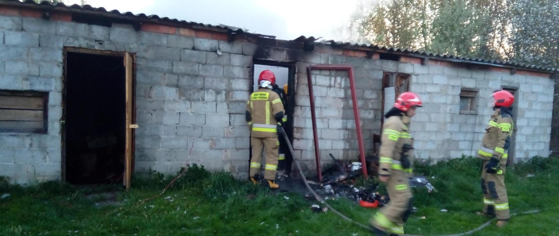 Na zdjęciu przedstawiono akcję ratowniczo-gaśniczą. 4 strażaków, w tym jeden podający wodę do wnętrza budynku przez otwór drzwiowy i jeden w aparacie ochrony dróg oddechowych wewnątrz obiektu, stoją na tle parterowego budynku murowanego gospodarczego.