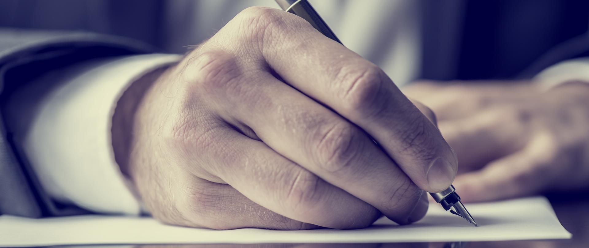 Man signing a document or writing correspondence with a close up view of his hand with the pen and sheet of notepaper on a desk top. With retro filter effect.
