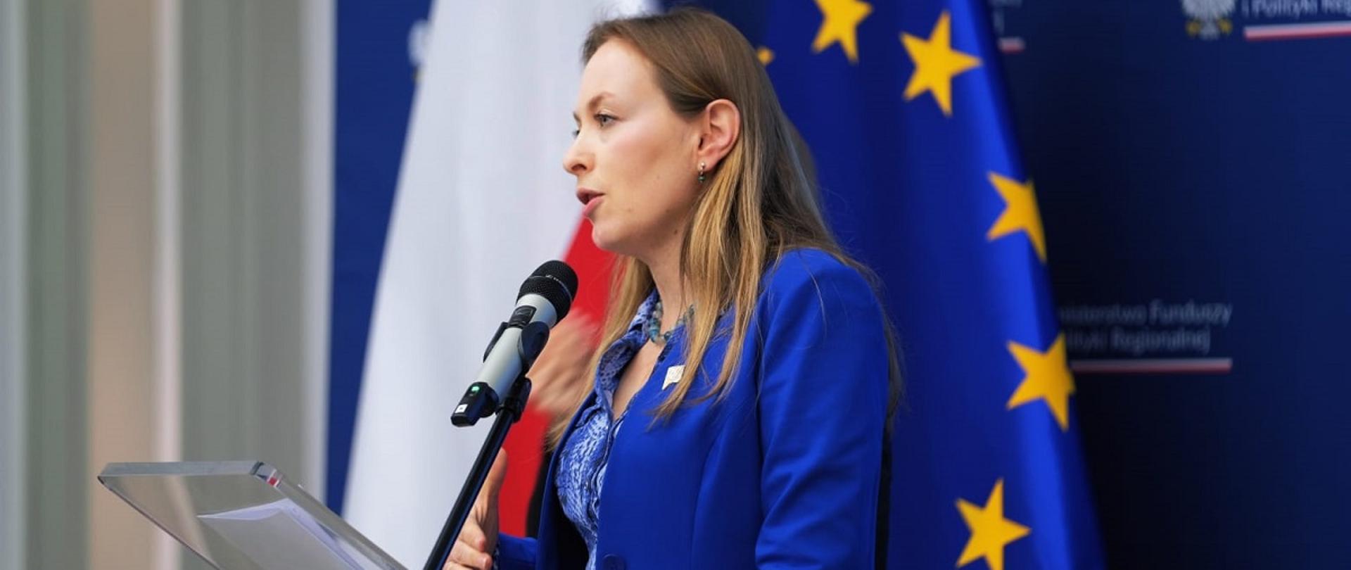 Minister funduszy i polityki regionalnej Katarzyna Pełczyńska-Nałęcz stoi przy pulpicie i przemawia do mikrofonu, za jej plecami flagi Polski i UE