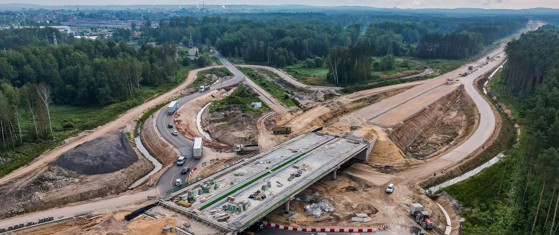Zdjęcie przedstawia lotniczy widok na duży plac budowy drogi. Widoczny jest częściowo ukończony wiadukt, a pod nim przebiega istniejąca droga. Na miejscu widoczne są maszyny i pojazdy budowlane Otaczający krajobraz obejmuje fragmenty obszarów zalesionych, a w oddali znajduje się zabudowany obszar miasta.