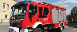 Na zdjęciu samochód strażacki widziany z przodu i boku. W tle budynek Komendy
