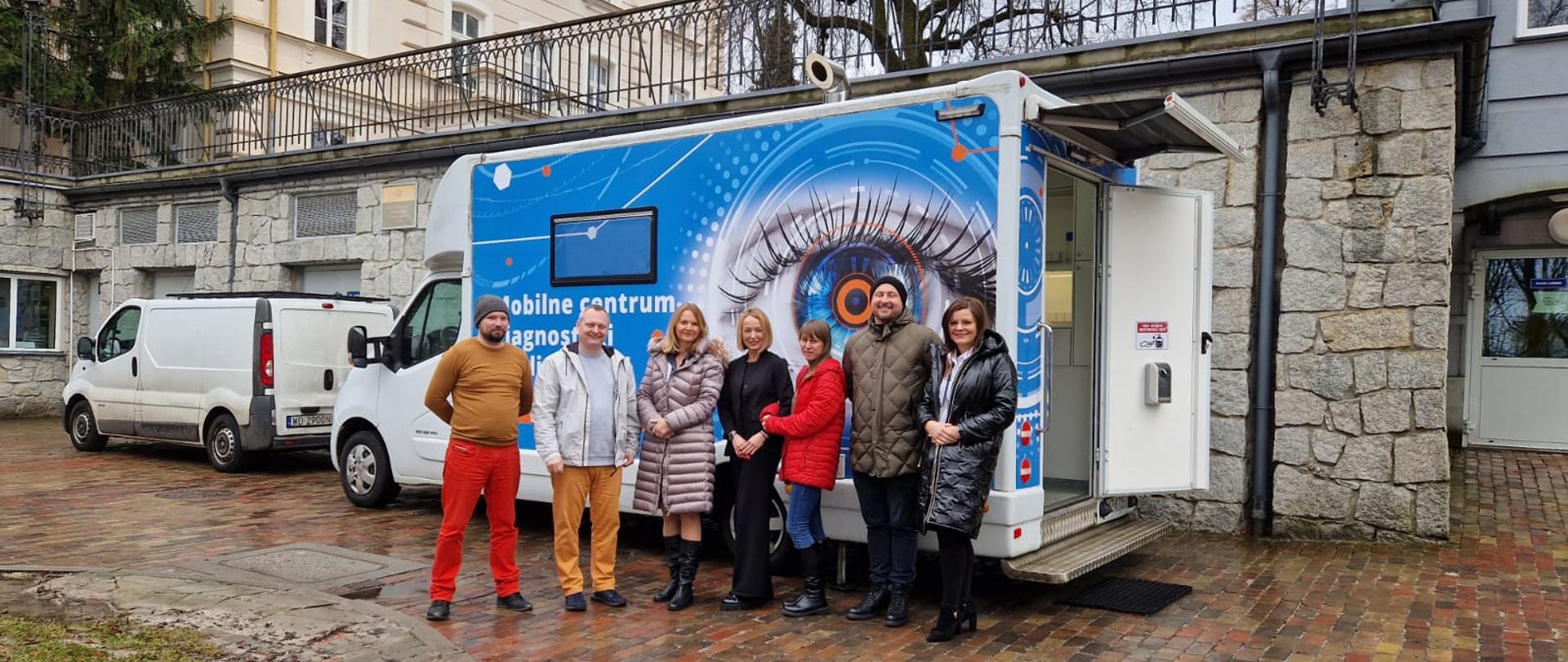 Grupa osób stoi w szeregu przed samochodem diagnostycznym Okobus. Pojazd jest zaparkowany przed zespołem budynków.