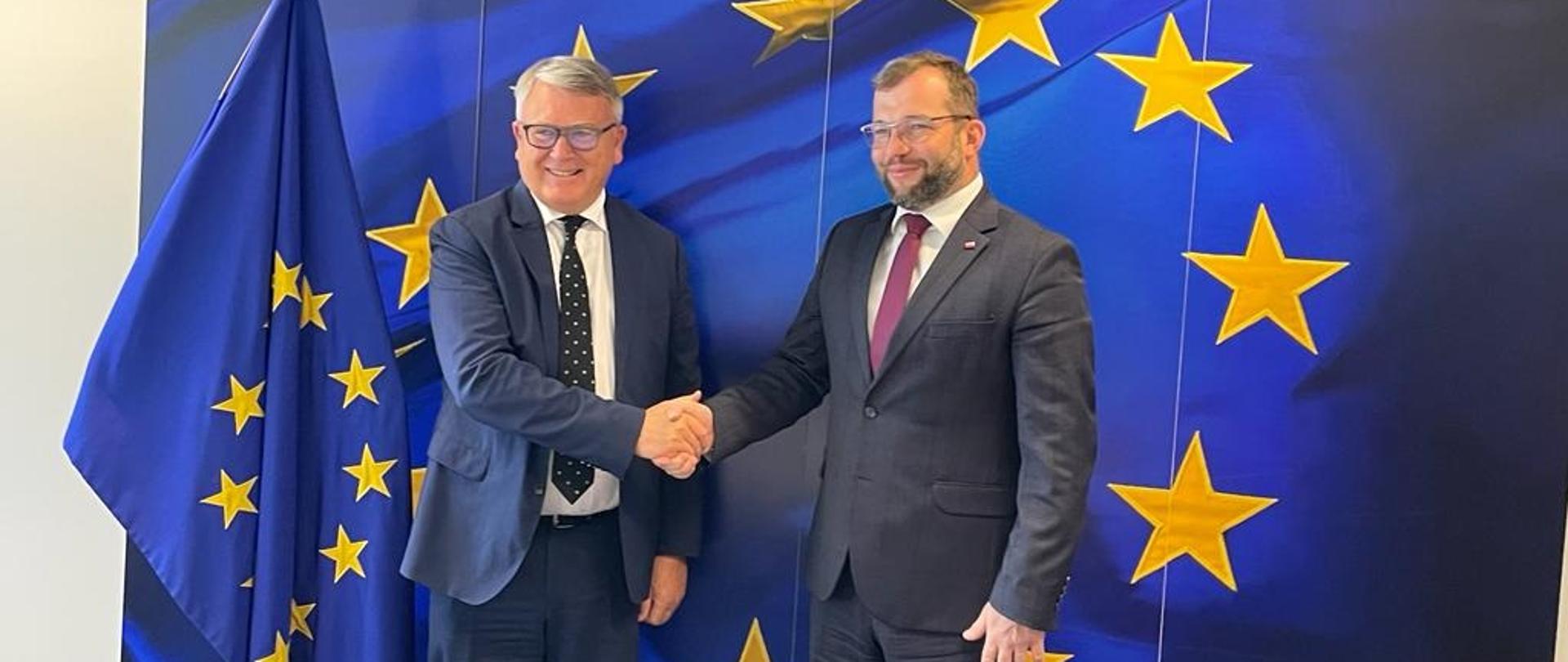Minister funduszy i polityki regionalnej Grzegorz Puda i komisarz do spraw miejsc pracy i praw socjalnych Nicolas Schmit stoją przy fladze UE i podają sobie dłonie na powitanie