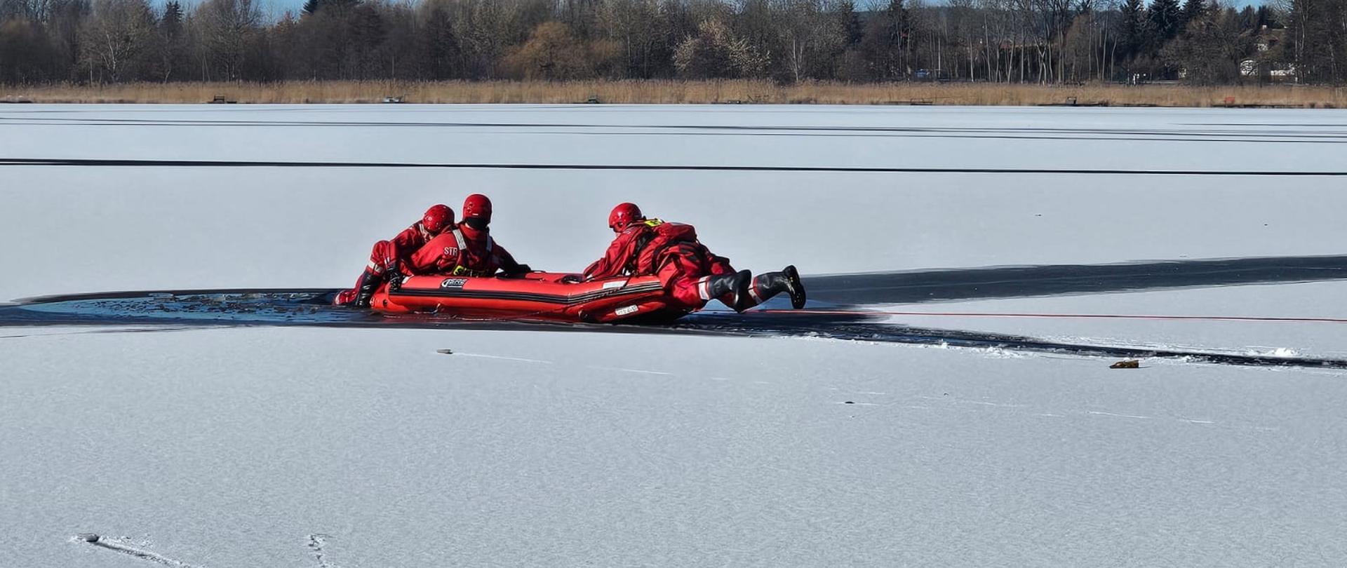 Zdjęcie przedstawia strażaków ćwiczących ratownictwo lodowe na jeziorze.
W tle jezioro o las.

