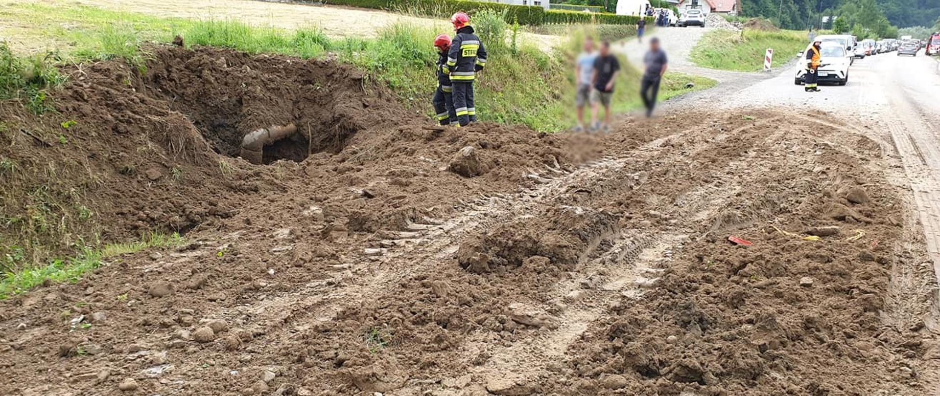 Błoto na drodze i widoczna wyrwa w ziemi obok której stoją strażacy.