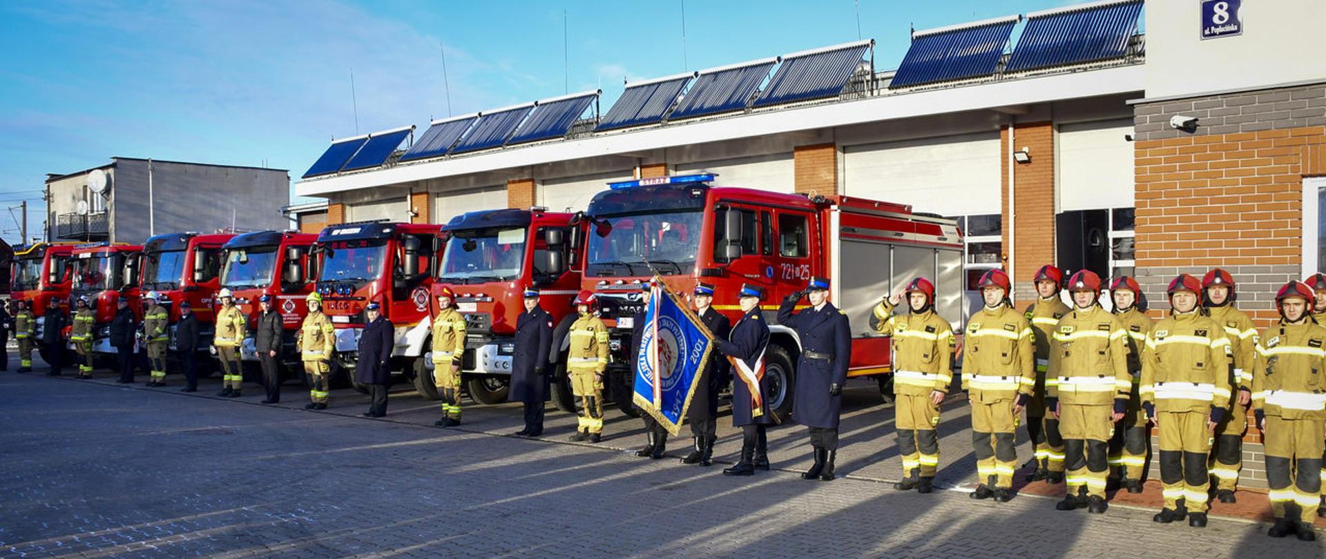 W rzędzie stoją strażacy. Prawej strony ubrani w mundury bojowe obok nich poczet sztandarowy z wysuniętym do przody sztandarem, z lewej strony strażacy w umundurowaniu wyjściowym na przemian ze strażakami w baniach bojowych. Za nimi stoją samochody pożarnicze.