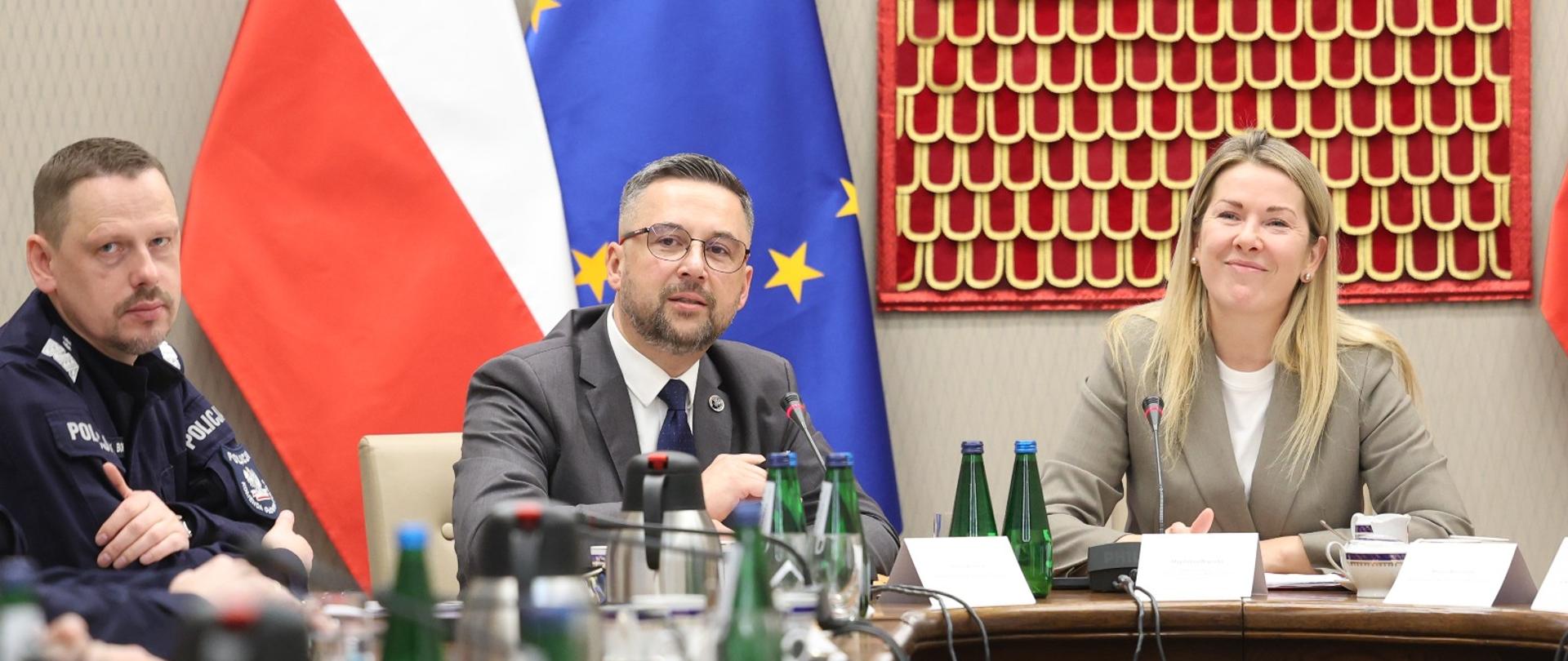 Za stołem siedzi minister Kulasek między mężczyzną w policyjnym mundurze i kobietą w jasnoszarej marynarce, z tyłu pod szarą ścianą flagi Polski i UE, na ścianie godło.