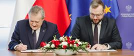 Minister kultury i dziedzictwa narodowego Piotr Gliński i minister funduszy i polityki regionalnej Grzegorz Puda podpisują porozumienie w sprawie realizacji wybranych zadań z programu Fundusze Europejskie na Infrastrukturę, Klimat, Środowisko 2021–2027, ministrowie siedzą przy stole i podpisują dokumenty, za ich plecami flagi Polski i UE