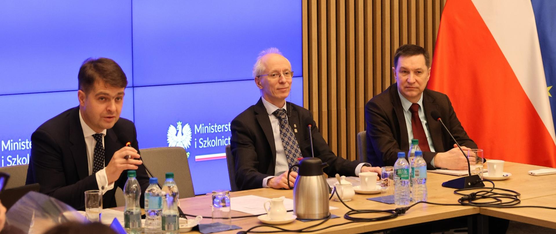 Widok na salę, przy stole siedzi trzech mężczyzn w garniturach, za nimi wbudowany w ścianę wielki ekran z napisem Ministerstwo Nauki i Szkolnictwa Wyższego.