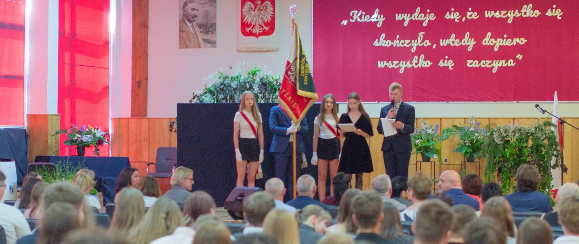 Widok zza pleców publiczności na scenę, na której stoi poczet sztandarowy oraz uczniowie prowadzący wydarzenie.