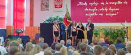 Widok zza pleców publiczności siedzącej w auli na scenę, na której stoi poczet sztandarowy z flagą oraz uczniowie prowadzący apel na tle dużej, czerwonej dekoracji z cytatem.