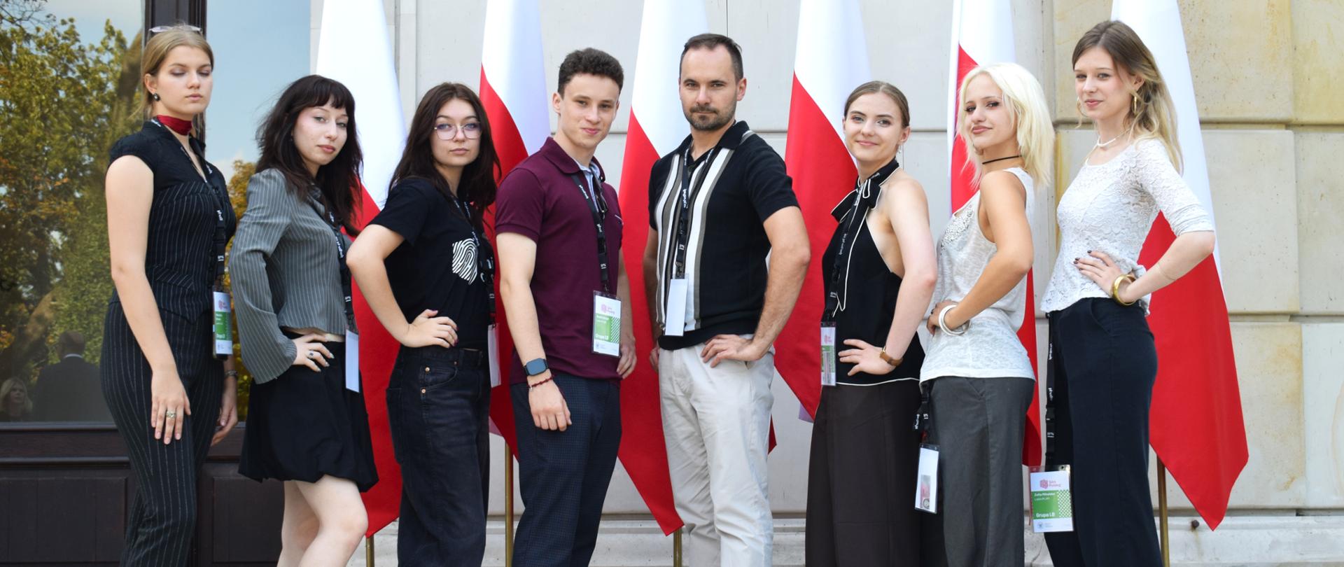 Siedem młodych osób i mężczyzna na dworze, przed budynkiem. Pozują na tle pięciu stojących za nimi flag polskich