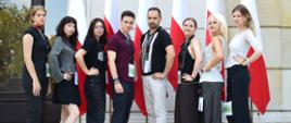 Siedem młodych osób i mężczyzna na dworze, przed budynkiem. Pozują na tle pięciu stojących za nimi flag polskich