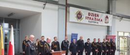 umundurowani strażacy stojący w szeregu w obiekcie garażu, po lewej stronie mównica z mikrofonem a przy niej przemawiający komendant powiatowy