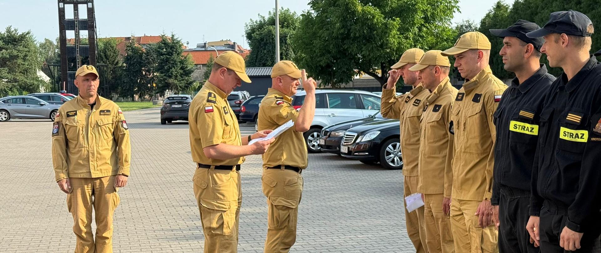 Komendant Powiatowy w asyście swojego zastępcy wręcza strażakom decyzje potwierdzające awans na wyższe stanowisko służbowe. Strażacy stoją w szyku. Ceremonia ma miejsce na zmianie służby w obecności dowódcy jrg.