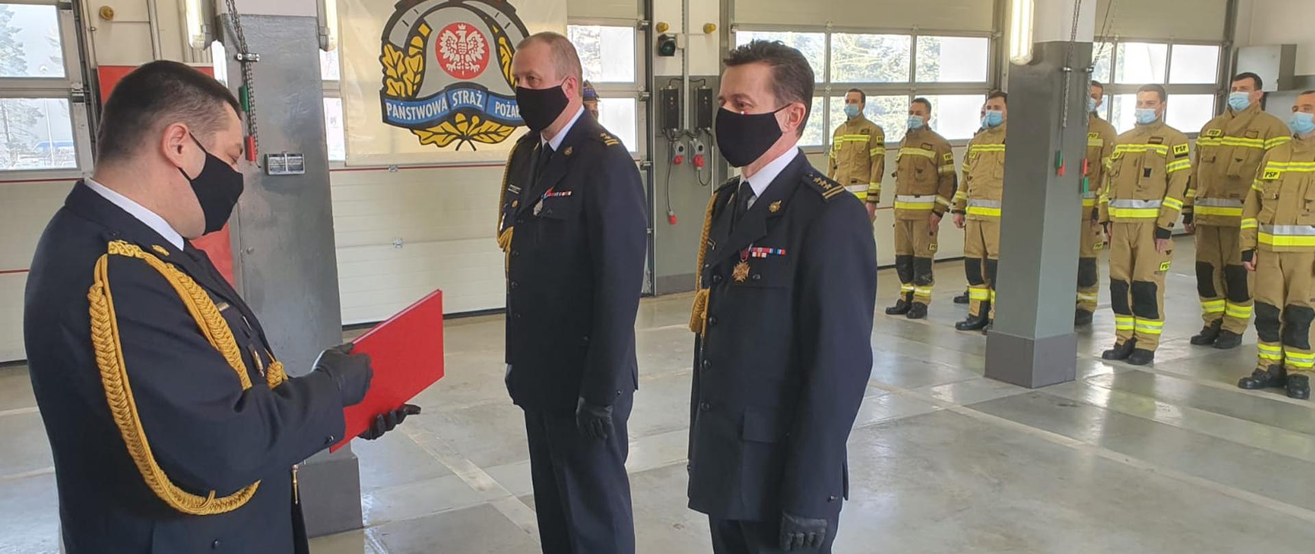 Zdjęcie przedstawia Śląskiego Komendanta Wojewódzkiego wręczającego akt odwołania st. bryg. Dariuszowi Kleszczowi w obecności bryg. Pawła Żaby. Komendanci w mundurze galowym obecni na garażu KM PSP w Jaworznie. W tle widoczne logo PSP oraz funkcjonariusze podziału bojowego w ubraniach specjalnych.