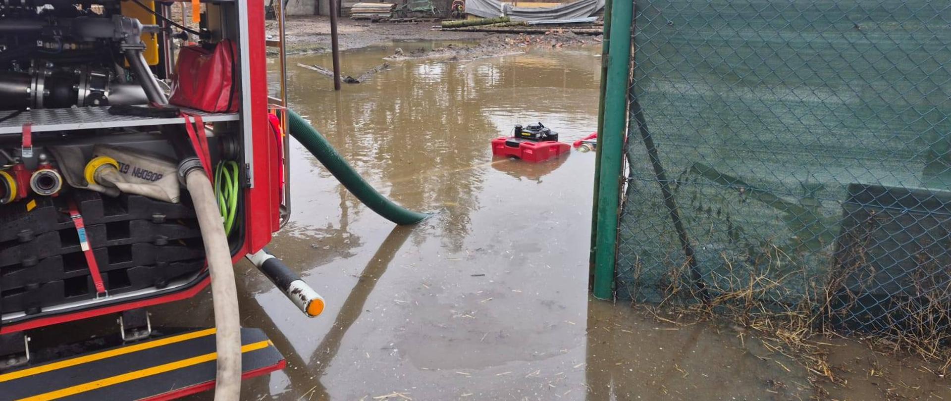 Zdjęcie przedstawia sprzęt ratowniczy i strażaków podczas wypompowywania wody w wyniku roztopów.
W tle zalane obiekty.
