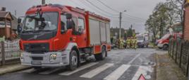 Na pierwszym planie stoi samochód pożarniczy PSP. Na lewym pasie widoczny trójkąt ostrzegawczy. W tle widać strażaków w ubraniach bojowych realizujących działania przy uszkodzonych pojazdach. Obok stoi karetka pogotowia oraz drugi pojazd pożarniczy