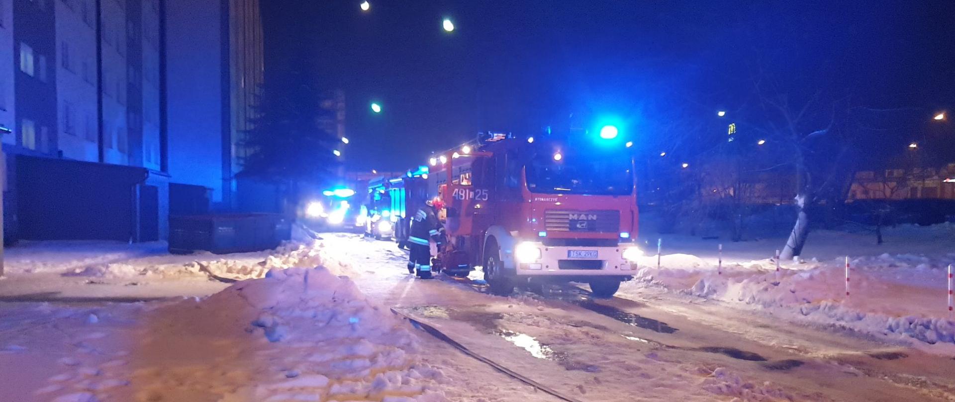 Na zdjęciu widać pojazdy straży pożarnej oraz ZRM z załączonymi sygnałami świetlnymi. Ratownik znajdujący się przy samochodzie przygotowuje sprzęt potrzebny do użycia przy pożarze piwnicy. 