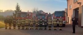 2. Na zdjęciu widoczni strażacy pełniący służbę w JRG Aleksandrów Kujawski. Funkcjonariusze w pełnych ubraniach specjalnych stoją w szeregu na tle pojazdów ratowniczych, z boku w mundurze wyjściowym widoczny Zastępca Komendanta PSP w Aleksandrowie Kujawskim. Strażacy zgromadzili się celem odsłuchania oraz uroczystego odśpiewania Hymnu narodowego.