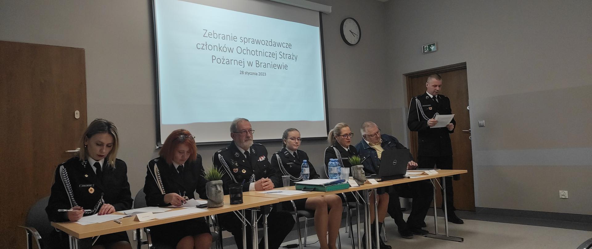Zdjęci e zrobione w pomieszczeniu. Za stolikami siedzi obok siebie sześciu strażaków ochotników, cztery kobiety i dwóch mężczyzn. Z prawej strony stoi strażak, czyta z kartki. Wszyscy w mundurach wyjściowych. Z tyłu na ścianie wyświetlana informacja z rzutnika.