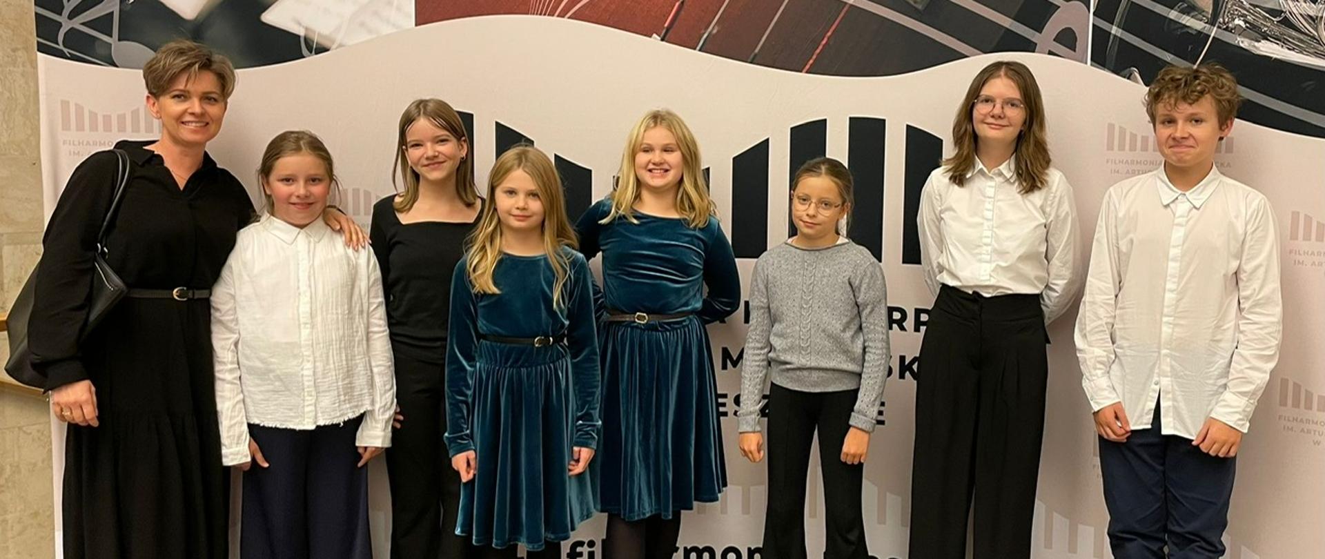 Grupa uczniów klasy fletu wraz z nauczycielką pozuje do wspólnego zdjęcia w holu Filharmonii Podkarpackiej. Wszyscy są elegancko ubrani, stoją na tle dekoracji z motywami muzycznymi przedstawiającymi instrumenty i nuty.