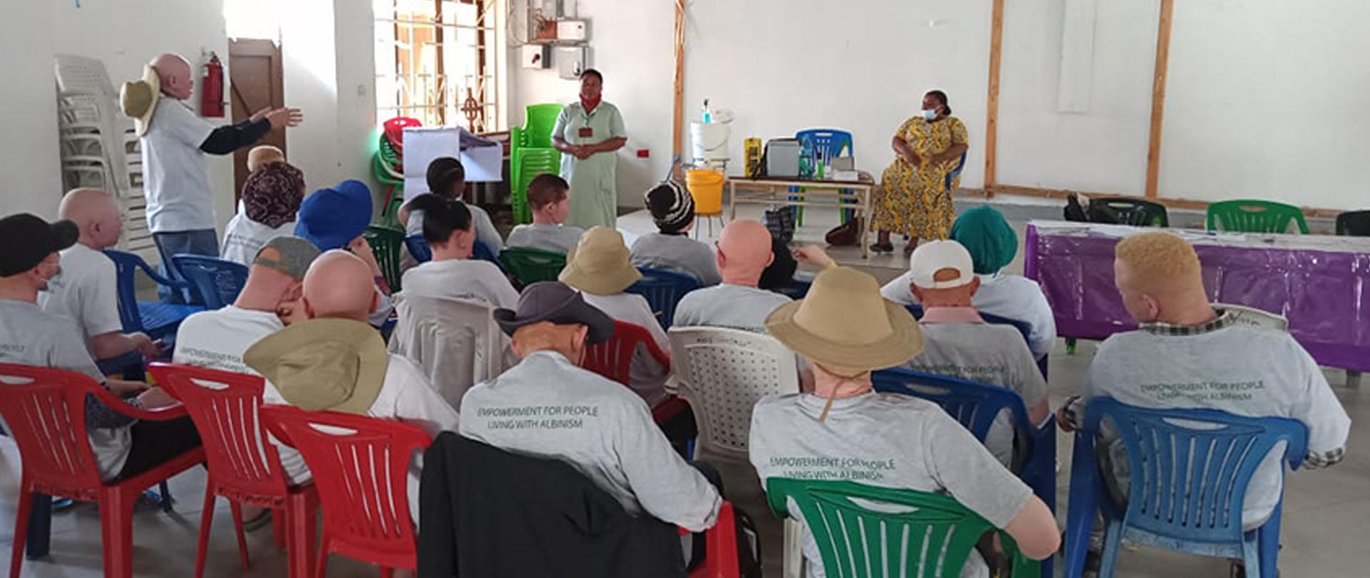 Grupa ludzi siedząca na plastikowych krzesłach. Jedna z nich, mężczyzna, stoi i rozmawia z kobietą, która stoi na podwyższeniu naprzeciw nich.