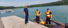 Na zdjęciu strażacy w łodzi oraz funkcjonariuszka Policji stojąca na pomoście 