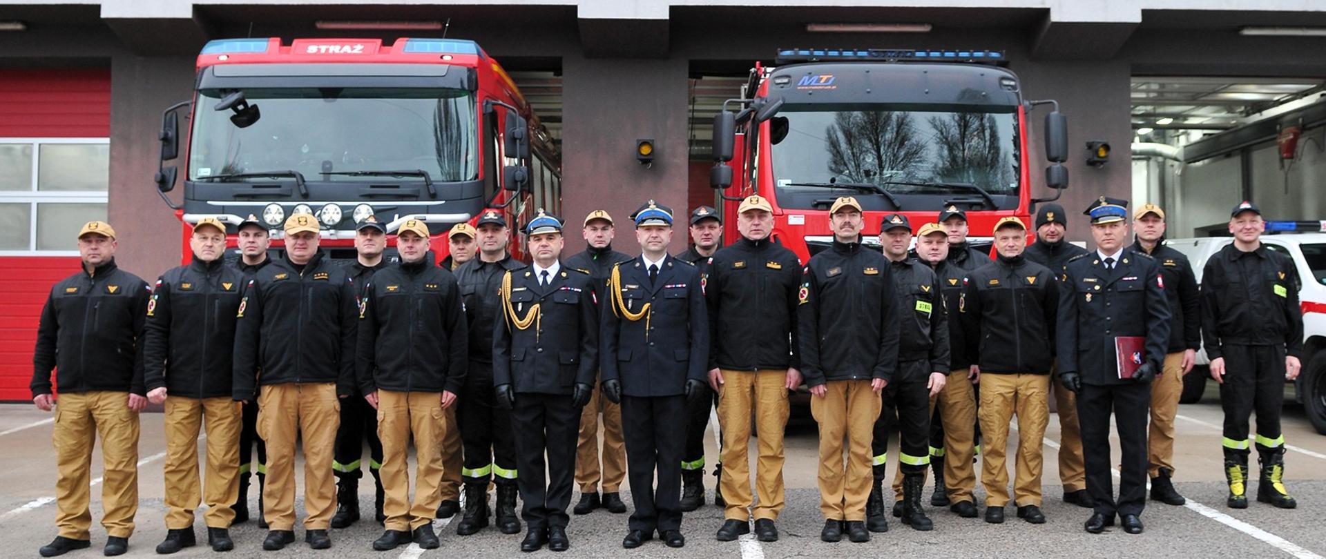 Zdjęcie przedstawia budynek Jednostki Ratowniczo-Gaśniczej Numer 2 z otwartymi bramami w których stoją samochody gaśnicze stanowiące tło dla strażaków. Wśród nich stoi odchodzący funkcjonariusz i zastępca komendanta miejskiego PSP w Kielcach.