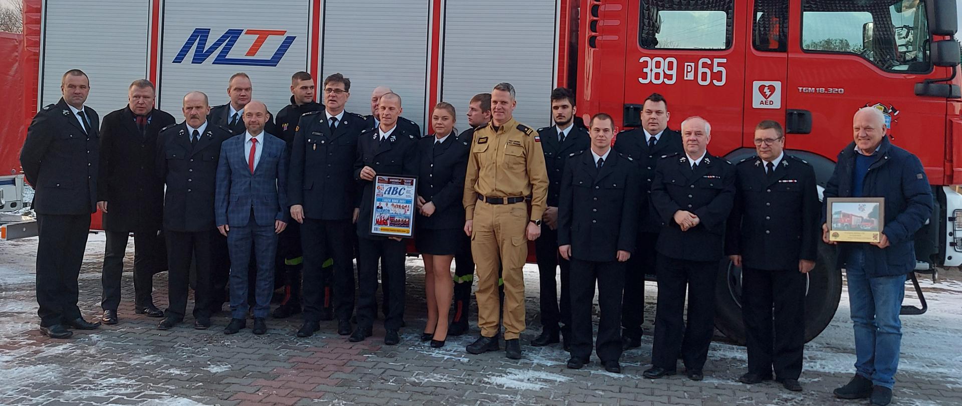 Strażacy w mudurach i osoby cywilne stoją w szeregu na tle samochodu strażackiego.