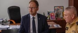 W pomieszczeniu z szarymi ścianami przy stole siedzą: prezydent miasta ubrany w granatowy garnitur oraz komendant w piaskowym mundurze. W rękach trzymają długopisy na stole leżą dokumenty.