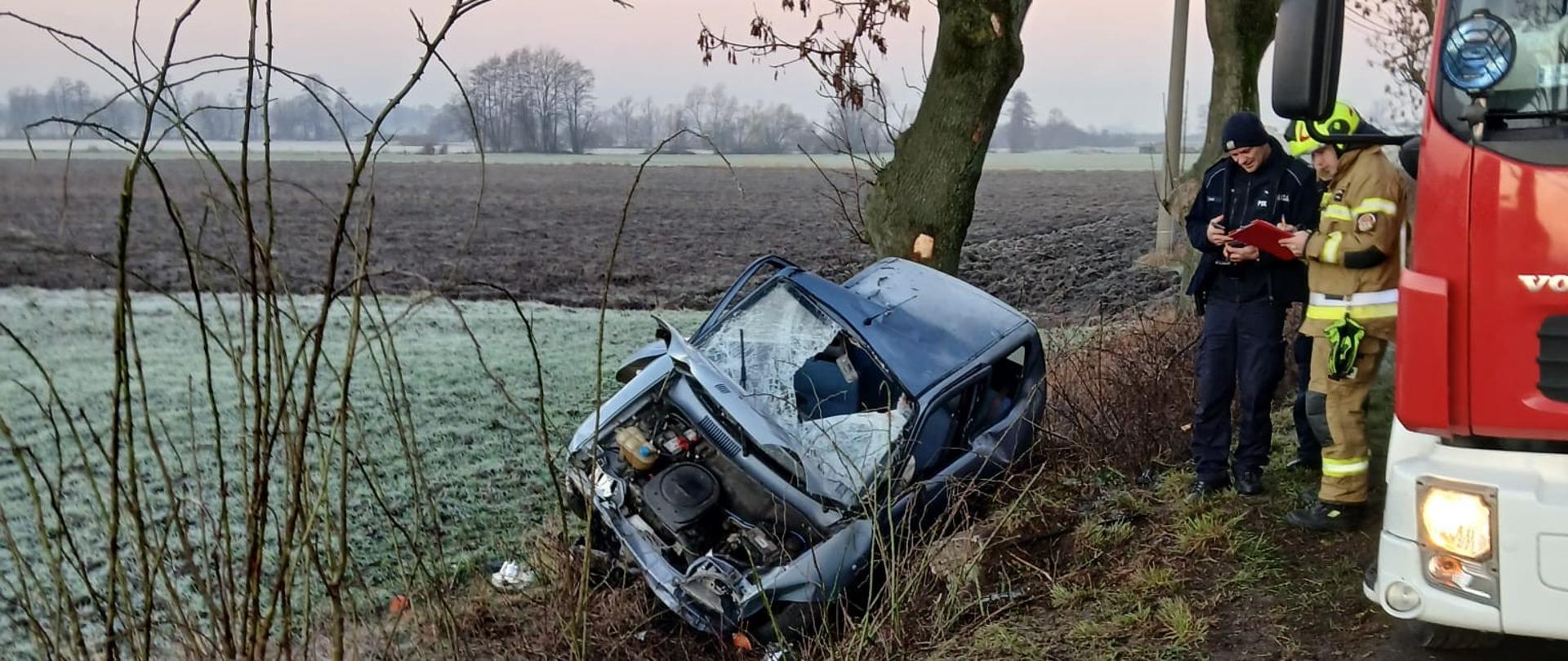 Na zdjęciu widać skutki wypadku drogowego. Samochód osobowy wypadł z jezdni i uderzył przodem w drzewo, po czym osunął się do rowu. Przód auta jest mocno zniszczony – maska jest zgnieciona, a elementy silnika są odsłonięte. Pojazd stoi przechylony wśród krzewów i trawy. Obok miejsca zdarzenia znajdują się służby ratunkowe. Widać strażaka w ubraniu ochronnym oraz funkcjonariusza policji, którzy stoją przy wozie strażackim zaparkowanym na poboczu. W tle rozciągają się pola pokryte szronem, drzewa oraz linie energetyczne.