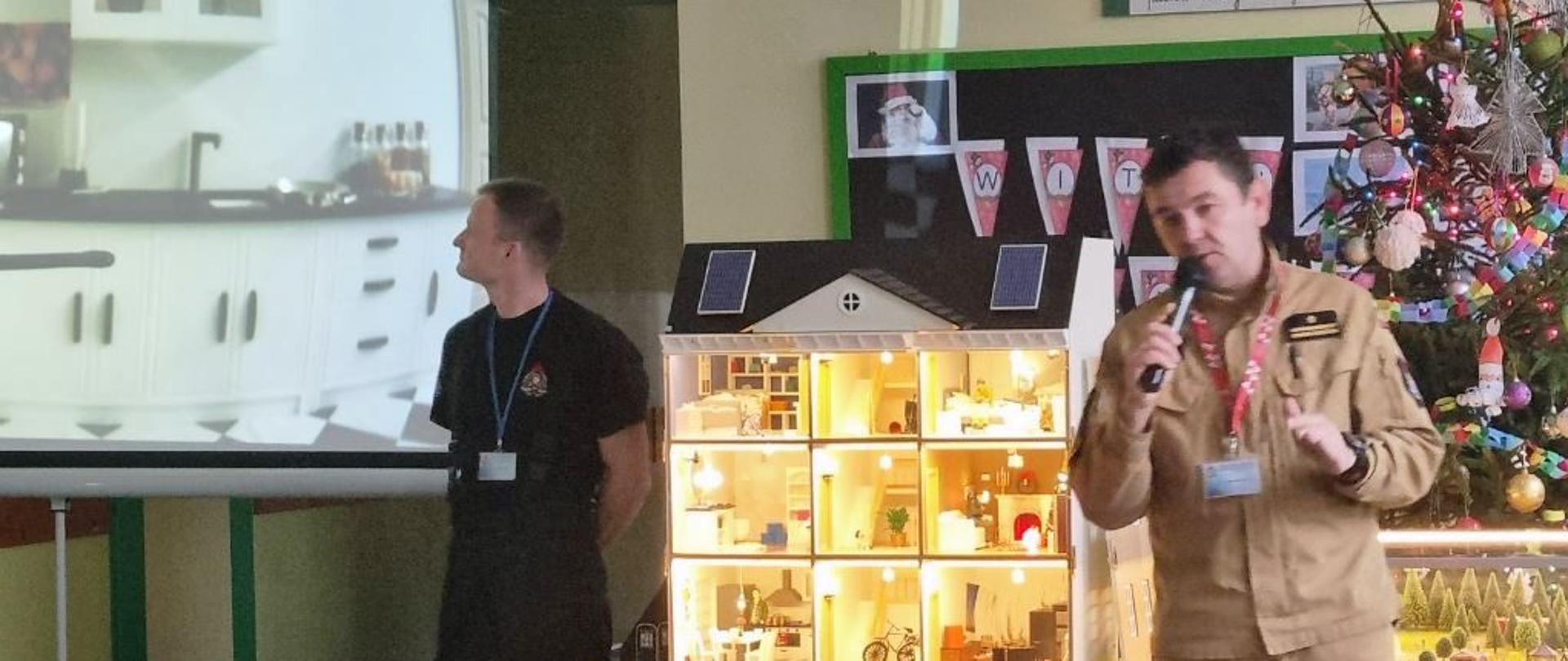 Strażak mówi trzymając mikrofon przy domku stoi funkcjonariusz Państwowej Straży Pożarnej przed nimi siedzą dzieci.