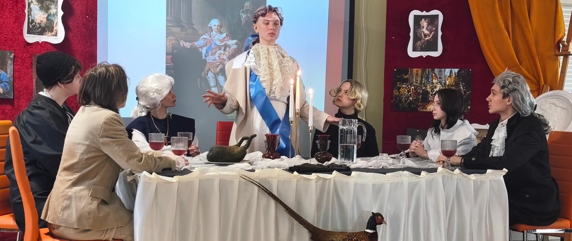 Scena została zaaranżowana jako uroczysta biesiada przy długim stole, nawiązująca do epoki nowożytnej (XVIII wiek). Uczestnicy są ubrani w stylizowane stroje historyczne: płaszcze, żaboty, peruki oraz eleganckie marynarki i suknie.
Na stole znajdują się: kielichy z czerwonym napojem, karafki, dzbanek, zapalone świece w lichtarzach, nadające scenie ceremonialny charakter.
W centrum stoi jedna osoba w jasnym stroju z niebieską szarfą.
Za uczestnikami wiszą dekoracyjne panele obite materiałem, z reprodukcjami malarstwa historycznego.
Na ekranie projekcyjnym widoczny jest portret króla Stanisława Augusta Poniatowskiego.
