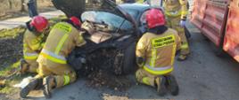 Kolizja drogowa-dachowanie audi. Droga gminna Rawicz-Przywsie. Na jezdni stoi auto osobowe uczestniczące w kolizji, które wcześniej leżało w przydrożnym rowie. Przy nim 4 strażaków odłączających akumulator. W tle drzewa.