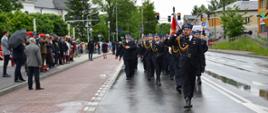 Fotografia przedstawia pododdział strażaków w mundurach wyjściowych maszerujący w cztergoszeregu po jednym pasie ulicy Siennickiej w Mińsku Mazowieckim podczas Powiatowego Dnia Strażaka. Pierwszy idzie dowódca uroczystości i oddaje honor stojącym na poboczu Ministrowi, Komendantom Państwowej Straży Pożarnej i pozostałym gościom, za nim idzie dowódca pododdziału, następnie sztandar, następnie pododdział w czteroszeregu 