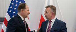 Minister Wieczorek stoi obok mężczyzny w garniturze, rozmawiają, za nimi pod białą ścianą flagi Polski i USA.
