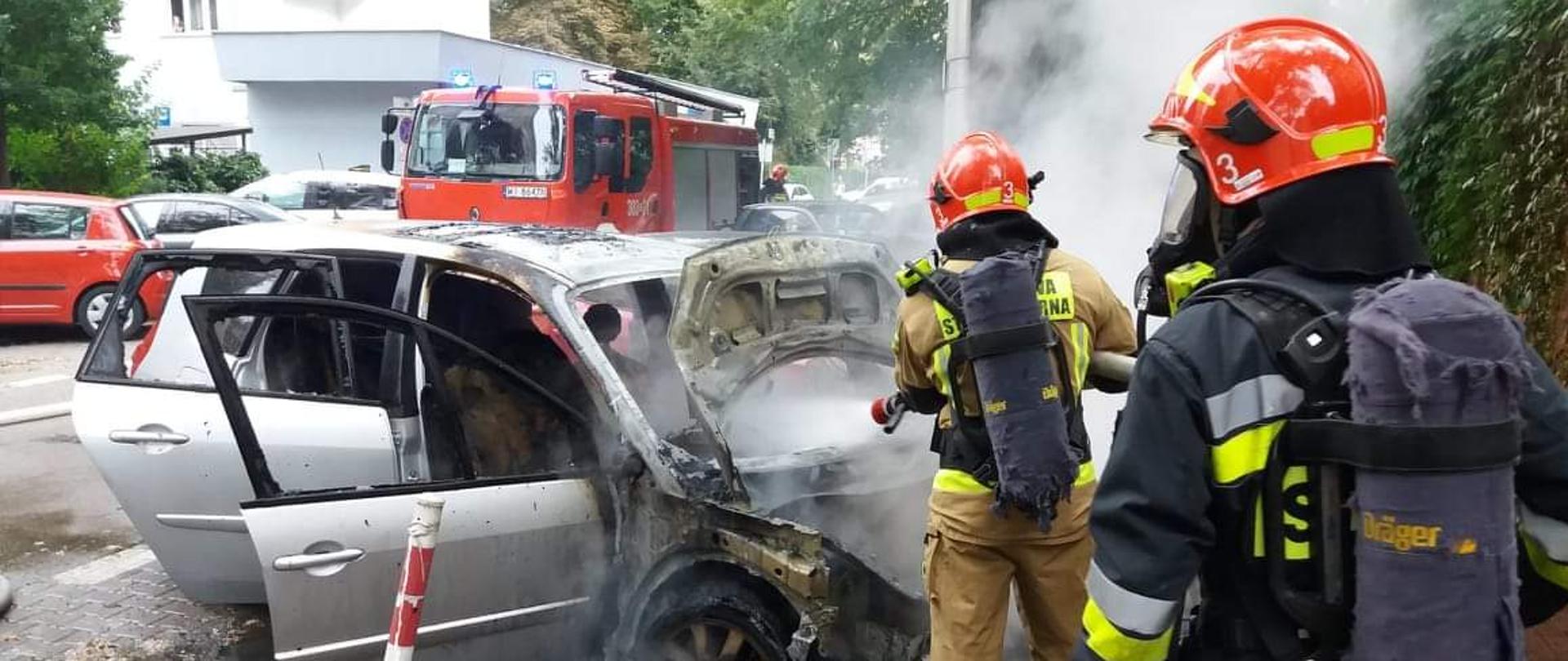 Na zdjęciu widać dwóch strażaków w aparatach z powietrzem gaszącym samochód osobowy. W tle widoczny samochód straży pożarnej.