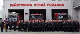 Widok ogólny uczestników zbiórki ustawionych na tle pojazdów pożarniczych i budynku JRG Andrychów.