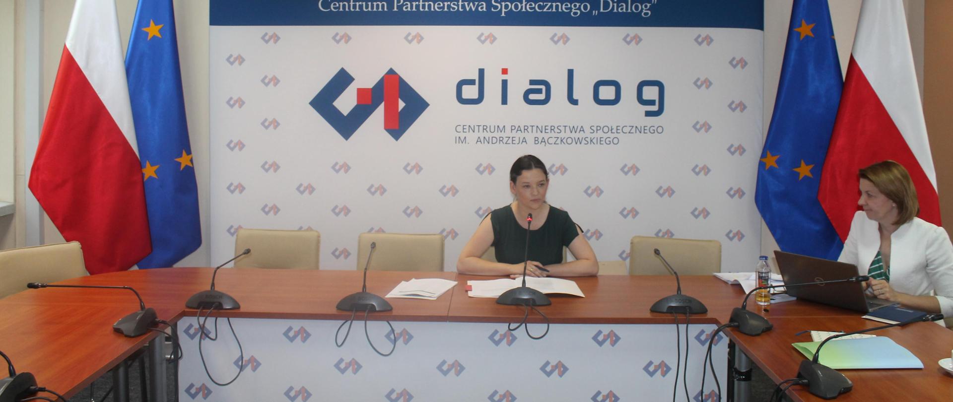 Na zdjęciu widnieje stół prezydialny za którym siedzą od lewej strony Katarzyna Nowakowska - Podsekretarz Stanu w Ministerstwie Rodziny, Pracy i Polityki Społeczne oraz Magdalena Fabisiak – Dyrektor Departamentu Pomocy Społecznej w MRPiPS. W tle siedzących znajduje się baner z napisem Rada Dialogu Społecznego a nad nim wisi godło Polski. Po obu stronach stołu widnieją dwie flagi: Polski i UE.