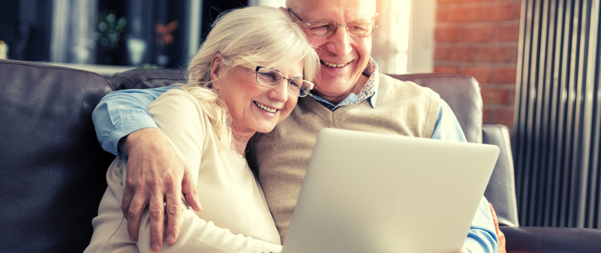 Dwoje uśmiechniętych starszych osób siedzi na kanapie, patrzą w laptopa. Mężczyzna obejmuje kobietę ramieniem. 