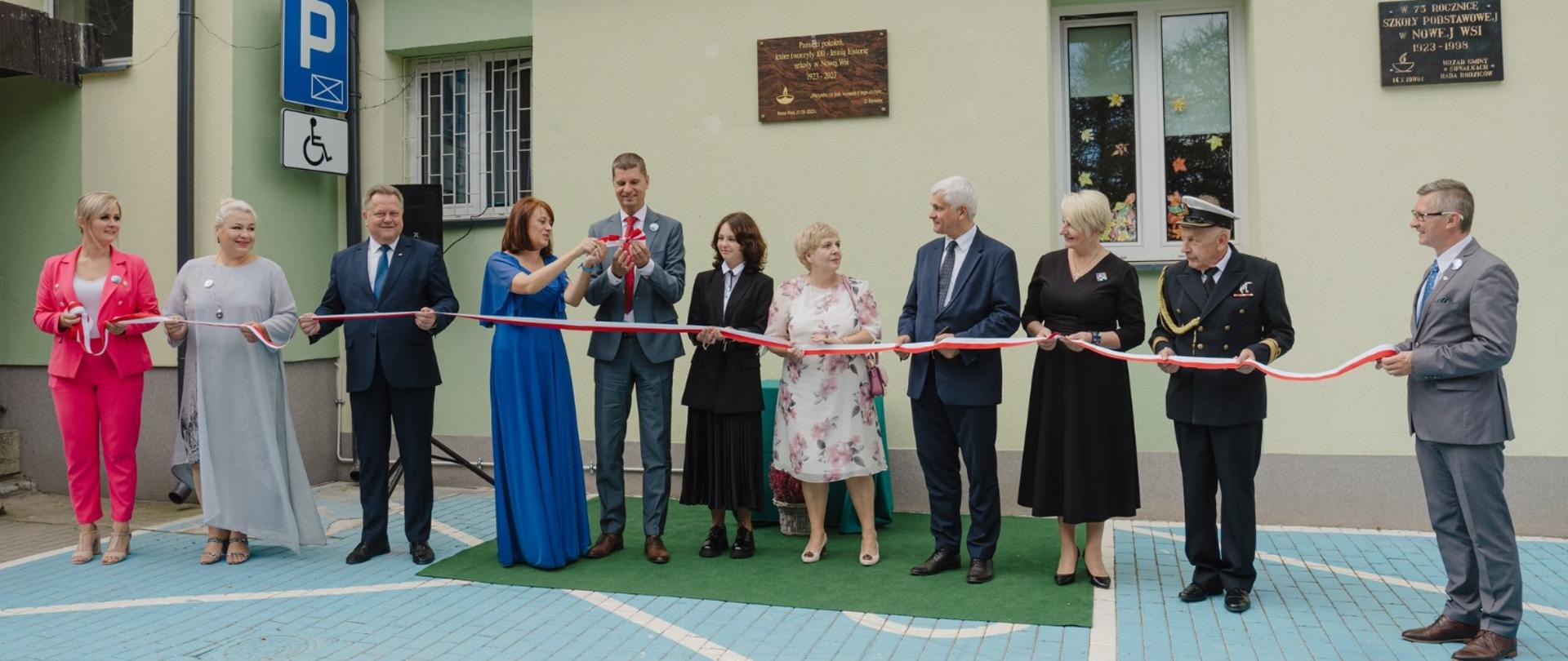 Przed jasnozieloną ścianą budynku stoi w rzędzie 11 osób, trzymają przed sobą biało-czerwoną wstążkę.