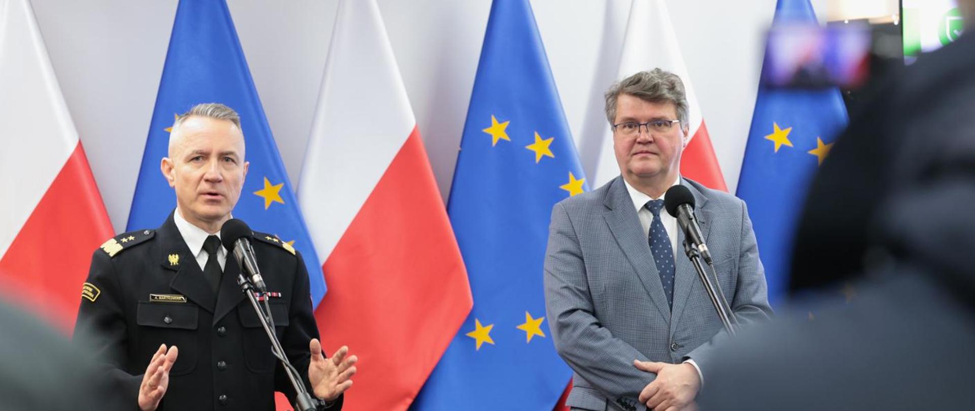 Na tle flag rzeczypospolitej Polskiej oraz Unii Europejskiej stoi dwóch mężczyzn. Z lewej strony w mundurze wyjściowym - komendant główny PSP, z prawej - wiceminister spraw wewnętrznych i administracji.