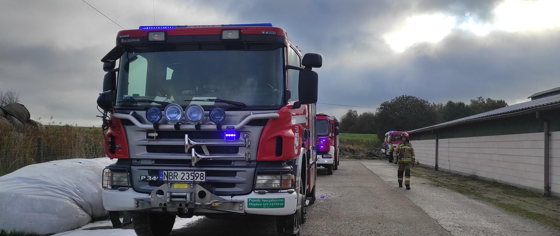Na zdjęciu z lewej strony stoją dwa samochody strażackie. Z prawej strony budynek gospodarczy, na końcu budynku za rogiem stoi kolejny czerwony samochód strażacki. Pochmurnie.
