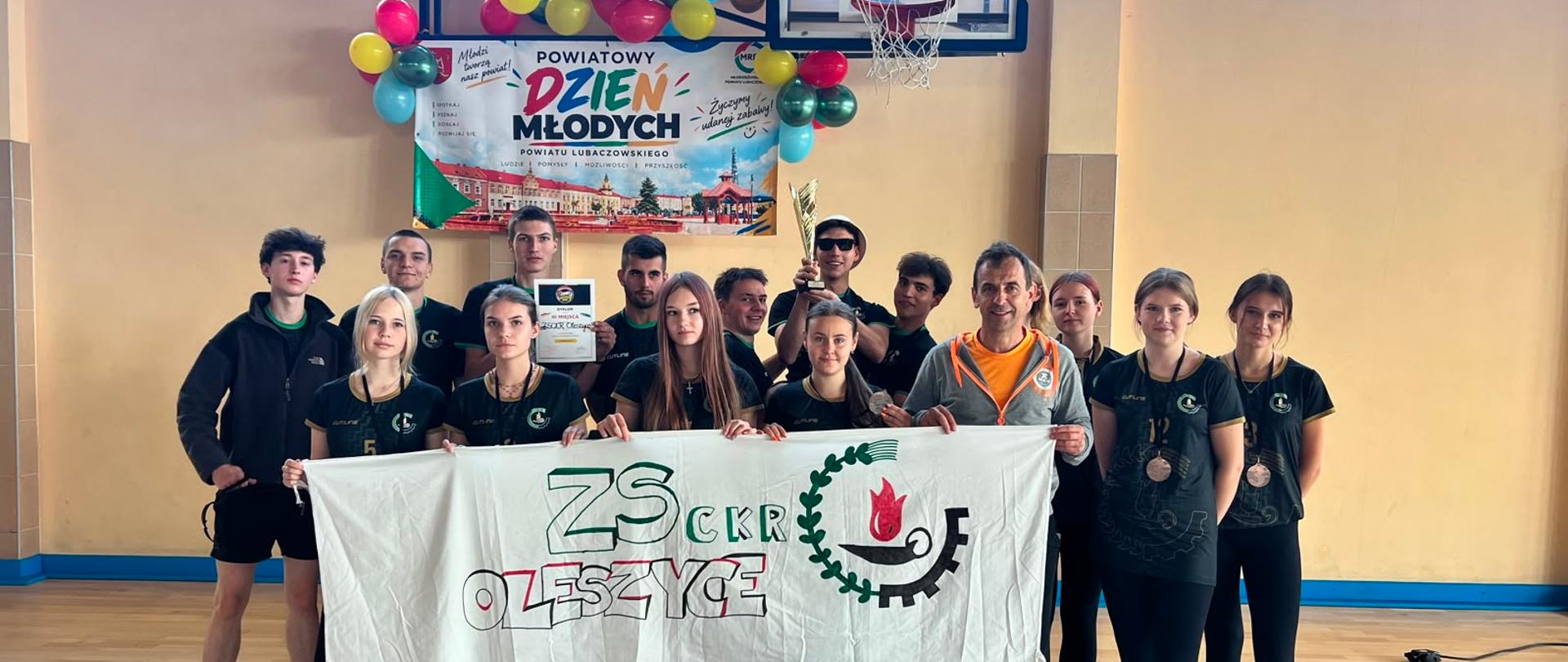 Grupa składa się z kilkunastu uczniów (zarówno dziewcząt, jak i chłopców) ubranych w sportowe stroje – większość ma na sobie ciemne koszulki z logo szkoły. Jeden z chłopców leży z przodu na parkiecie, trzymając upominek. W tłumie widoczny jest również dorosły mężczyzna w pomarańczowo-szarej bluzie sportowej, prawdopodobnie opiekun lub nauczyciel.